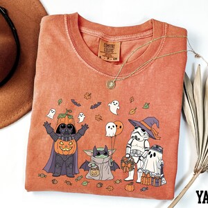 a Halloween-themed t-shirt with a design of various characters, including Darth Vader, Baby Yoda, Stormtrooper, and Ghost Batter, along with Halloween-related elements such as pumpkins, bats, and leaves.