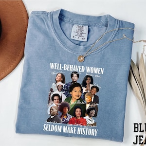 A blue t-shirt with a graphic design featuring a collage of diverse women's faces, along with the text "WELL-BEHAVED WOMEN" and "SEldom MAKE HISTORY".