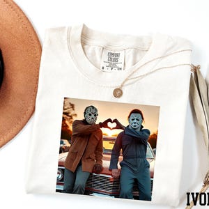 a white t-shirt with a printed photo of two people posing with their hands in a heart shape, along with a necklace and a hat.