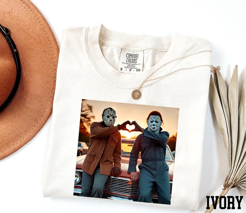 a white t-shirt with a printed photo of two people posing with their hands in a heart shape, along with a necklace and a hat.