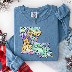 a blue long-sleeved t-shirt with a colorful graphic of Louisiana, a state in the United States, along with various decorative elements such as stars and a crown. The t-shirt is laid out on a white surface, accompanied by a pair of jeans and some decorative accessories.