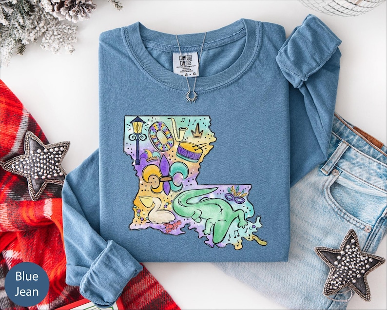 a blue long-sleeved t-shirt with a colorful graphic of Louisiana, a state in the United States, along with various decorative elements such as stars and a crown. The t-shirt is laid out on a white surface, accompanied by a pair of jeans and some decorative accessories.
