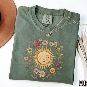a green t-shirt with a sun design, a brown hat, and a necklace, all placed on a white surface.