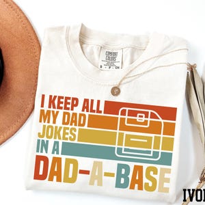 May include: A white t-shirt with a retro-style graphic that says "I keep all my dad jokes in a dad-a-base". The graphic features a floppy disk and colourful stripes. The shirt is ivory.