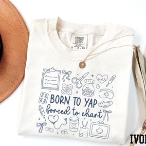 May include: Ivory-colored t-shirt with the text "BORN TO YAP forced to chart" and medical-themed illustrations in navy blue. The shirt has a crew neck and short sleeves. A gold necklace is draped across the shirt.