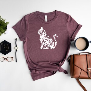 May include: A mauve t-shirt with a white silhouette of a cat with a damask pattern. The cat is sitting with its tail curled around its body. The t-shirt is laid flat on a white surface with a fern plant, a pair of glasses, a watch, a small black tray with earrings, a brown leather purse, and a cup of coffee.