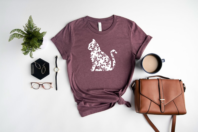 May include: A mauve t-shirt with a white silhouette of a cat with a damask pattern. The cat is sitting with its tail curled around its body. The t-shirt is laid flat on a white surface with a fern plant, a pair of glasses, a watch, a small black tray with earrings, a brown leather purse, and a cup of coffee.