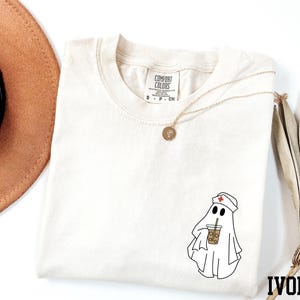 a white t-shirt with a graphic of a ghost wearing a nurse's hat and holding a drink, along with a necklace and a brown hat.