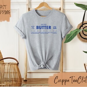 Puede incluir: Camiseta gris con un gr&aacute;fico azul de una etiqueta de mantequilla que dice "Salted Butter Grade AA" e incluye una tabla para medir la mantequilla en cucharadas y tazas.