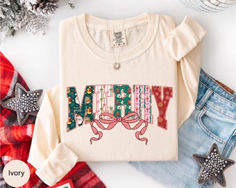 May include: Ivory-colored long-sleeve shirt with the word "MERRY" in a festive design. The letters are filled with Christmas-themed patterns and a red bow. The shirt is laid flat on a white surface.