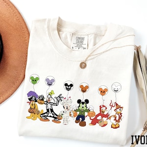 A white t-shirt with a graphic design of various Disney characters, including Mickey Mouse, Donald Duck, Goofy, and others, along with balloons. The t-shirt is placed on a white surface, and there is a brown hat and a fan nearby.