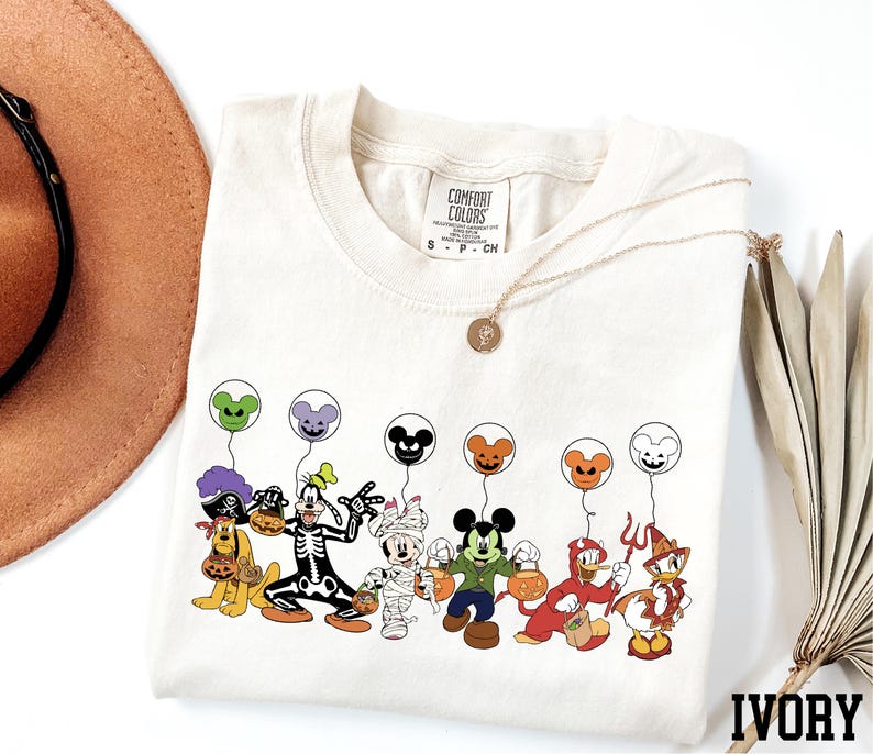 A white t-shirt with a graphic design of various Disney characters, including Mickey Mouse, Donald Duck, Goofy, and others, along with balloons. The t-shirt is placed on a white surface, and there is a brown hat and a fan nearby.
