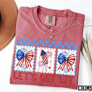a red t-shirt with a patriotic design, including American flags and fireworks, along with the text "Let's Get Lit" printed on it.
