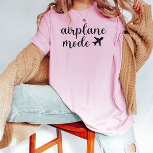 Puede incluir: Una camiseta rosa con el texto "airplane mode" y un peque&ntilde;o avi&oacute;n impreso en la parte delantera.