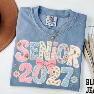 A blue denim t-shirt with the text "SENIOR 2027" printed on it, along with floral and polka dot designs. The t-shirt is folded and placed on a white surface, accompanied by a brown hat, a necklace, and some leaves.