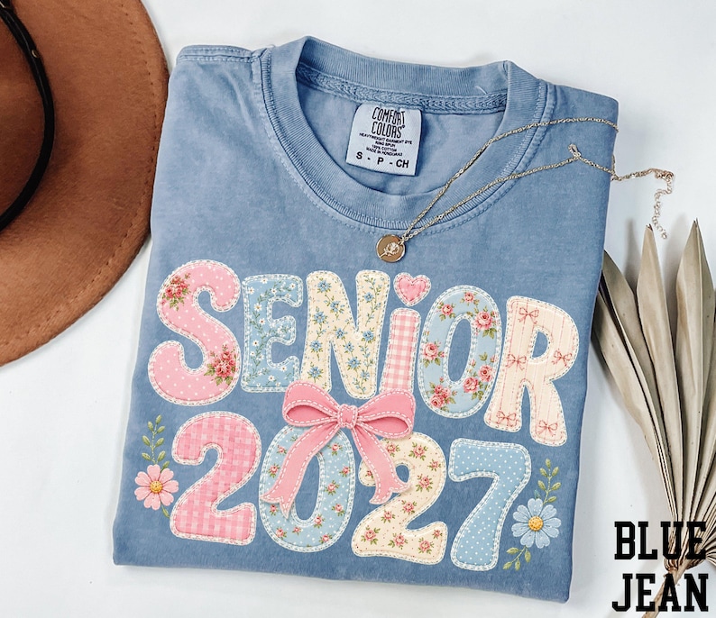 A blue denim t-shirt with the text "SENIOR 2027" printed on it, along with floral and polka dot designs. The t-shirt is folded and placed on a white surface, accompanied by a brown hat, a necklace, and some leaves.