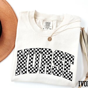 a white t-shirt with the word "NURSE" printed in black and white checkered letters, along with a brown hat and a necklace.