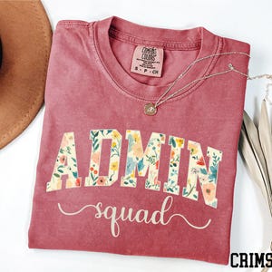 May include: Crimson-colored t-shirt with the word "ADMIN" in a floral design and the word "squad" in a cursive font. The shirt has a crew neck and short sleeves. A silver necklace with a pendant is on the shirt.