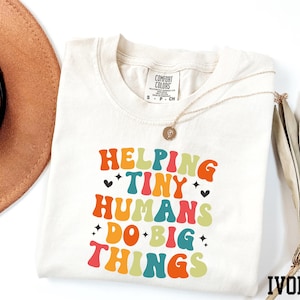 May include: Ivory-coloured t-shirt with the text "Helping Tiny Humans Do Big Things" in a retro font. The shirt has a crew neck and short sleeves. A gold necklace is draped across the shirt.