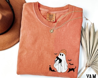 May include: A coral-coloured t-shirt with a crew neck and a graphic of a ghost wearing an orange beanie and holding a black cat. Two additional black cats and three bats are also featured. A gold necklace and a brown hat are also visible.