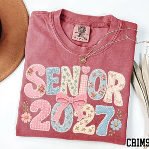 a pink t-shirt with the text "SENIOR 2027" printed on it, along with floral and bow designs. The t-shirt is folded and placed on a white surface, accompanied by a brown hat, a necklace, and some leaves.