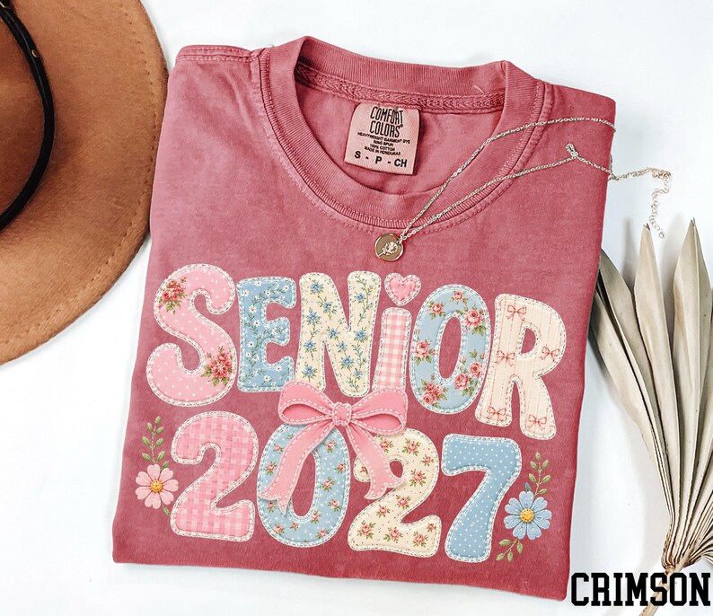 a pink t-shirt with the text "SENIOR 2027" printed on it, along with floral and bow designs. The t-shirt is folded and placed on a white surface, accompanied by a brown hat, a necklace, and some leaves.