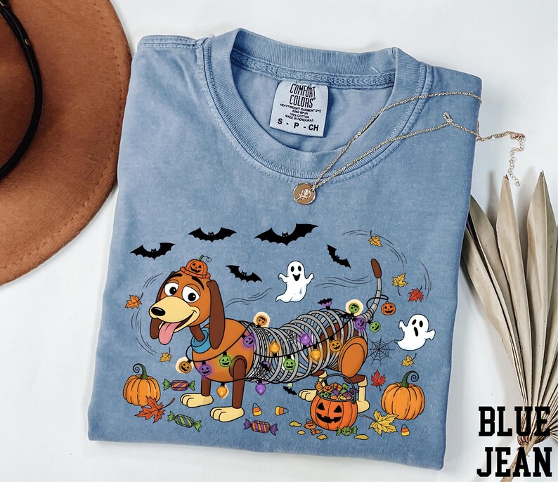 A blue t-shirt with a Halloween-themed design, including a cartoon dog, bats, a ghost, and pumpkins, along with a necklace and a hat.