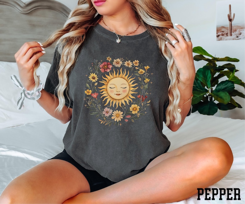 A woman with long, blonde hair is sitting on a bed, wearing a gray t-shirt with a sun design and surrounded by potted plants.