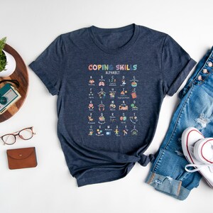 May include: A navy blue t-shirt with a graphic of a colorful alphabet chart that says "Coping Skills Alphabet". The chart features icons representing different coping skills for each letter of the alphabet.