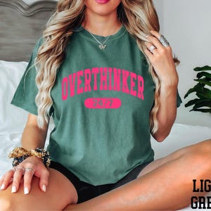 A woman wearing a green t-shirt with the text "OVERTHINKER" and the number "24/7" printed on it, sitting on a bed with a potted plant in the background.