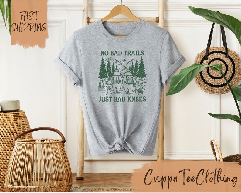 Może przedstawiać: Szara koszulka z zielonym nadrukiem przedstawiającym dw&oacute;ch niedźwiedzi wędrujących po lesie. Na koszulce widnieje napis "No Bad Trails Just Bad Knees".
