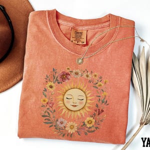 a pink t-shirt with a sun design, a brown hat, and a necklace, all arranged on a white background.