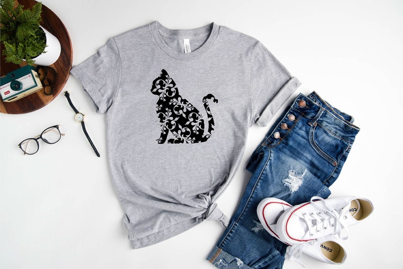 May include: A gray t-shirt with a black silhouette of a cat with a floral pattern. The cat is sitting with its tail curled around its body. The t-shirt is laid out on a white surface with a pair of blue jeans and white sneakers.