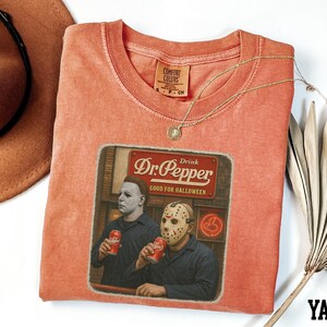 a red t-shirt with a graphic design depicting two men wearing masks, one holding a can of Dr. Pepper. The t-shirt is placed on a white surface, with a brown hat and a fan visible in the background.