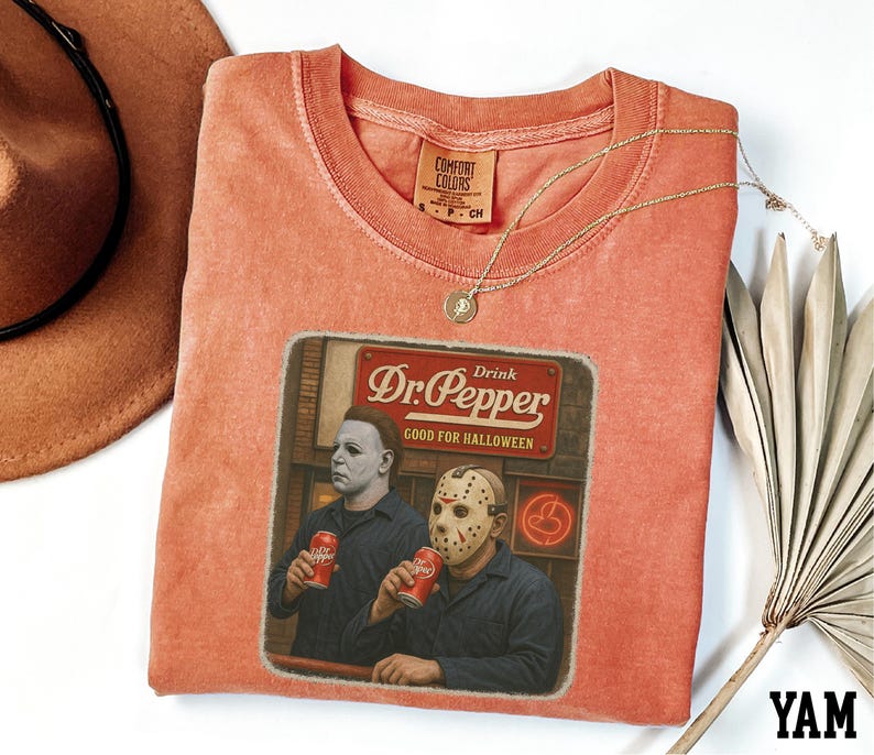 a red t-shirt with a graphic design depicting two men wearing masks, one holding a can of Dr. Pepper. The t-shirt is placed on a white surface, with a brown hat and a fan visible in the background.