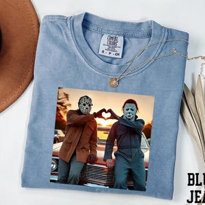 A blue t-shirt with a printed image of two people wearing masks, one with a pumpkin head and the other with a mask resembling a skeleton, posing in front of a car.