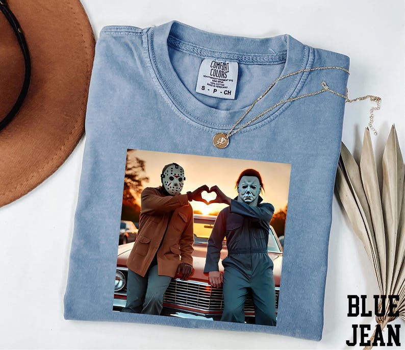 A blue t-shirt with a printed image of two people wearing masks, one with a pumpkin head and the other with a mask resembling a skeleton, posing in front of a car.