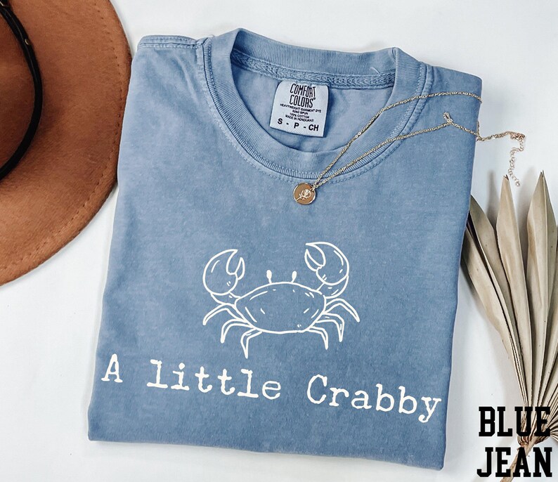 A blue t-shirt with a white crab design and the text "A little Crabby" printed on it, along with a necklace and a brown hat.