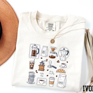 a white t-shirt with a graphic design of various coffee-related items, such as a coffee maker, coffee cup, and coffee beans, along with the text "all day coffee club". The t-shirt is laid out on a white background, and there is a brown hat and a fan visible in the background.