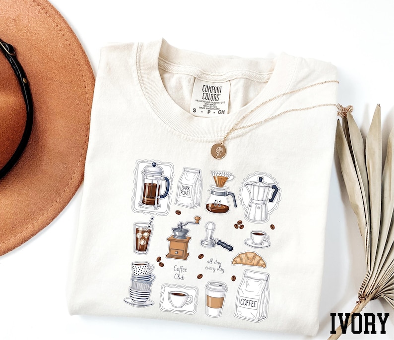 a white t-shirt with a graphic design of various coffee-related items, such as a coffee maker, coffee cup, and coffee beans, along with the text "all day coffee club". The t-shirt is laid out on a white background, and there is a brown hat and a fan visible in the background.