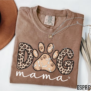 May include: Espresso-colored t-shirt with a dog paw print design. The design features the word "DOG" in a leopard print pattern, a paw print with bones, and the word "mama" in white script. A silver necklace with a pendant is on the shirt.
