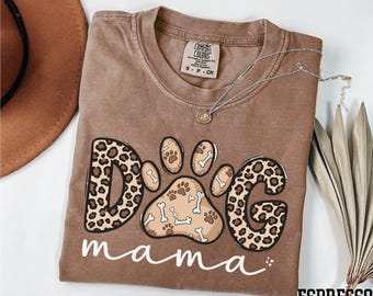 May include: Espresso-colored t-shirt with a dog paw print design. The design features the word "DOG" in a leopard print pattern, a paw print with bones, and the word "mama" in white script. A silver necklace with a pendant is on the shirt.