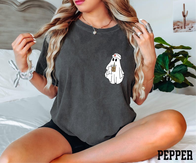 A woman with long blonde hair is sitting on a bed, wearing a black t-shirt with a ghost graphic and holding a drink.