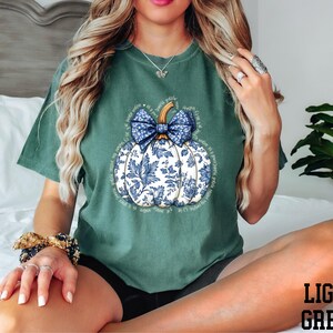 A woman wearing a green t-shirt with a blue and white pumpkin graphic, sitting on a bed with a potted plant in the background.