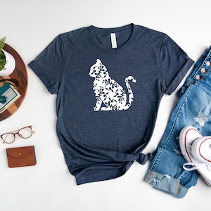 May include: A navy blue t-shirt with a white silhouette of a cat made of a floral pattern. The t-shirt is laid flat on a white surface with a pair of jeans and white sneakers.