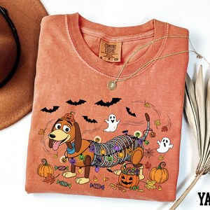 a pink t-shirt with a Halloween-themed design, including a cartoon dog, bats, a ghost, and pumpkins.