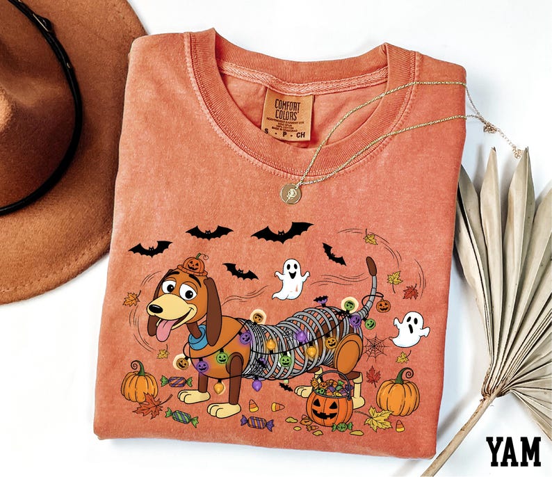 a pink t-shirt with a Halloween-themed design, including a cartoon dog, bats, a ghost, and pumpkins.