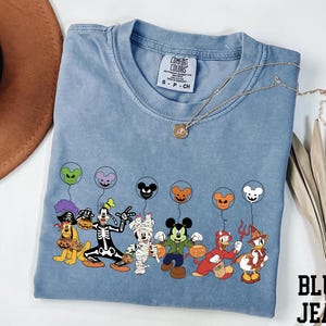 A blue t-shirt with a graphic of various Disney characters, including Mickey Mouse, Donald Duck, Goofy, and others, along with balloons. The t-shirt is placed on a white surface, and there is a brown hat and some leaves nearby.