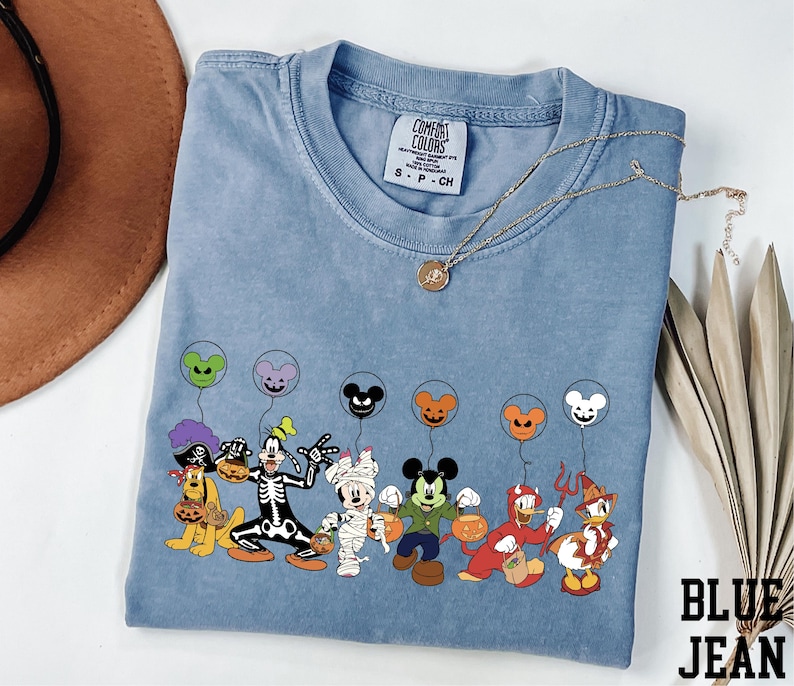 A blue t-shirt with a graphic of various Disney characters, including Mickey Mouse, Donald Duck, Goofy, and others, along with balloons. The t-shirt is placed on a white surface, and there is a brown hat and some leaves nearby.