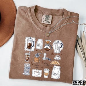 a brown t-shirt with a graphic design depicting various coffee-related items such as a coffee maker, coffee cup, and coffee beans.